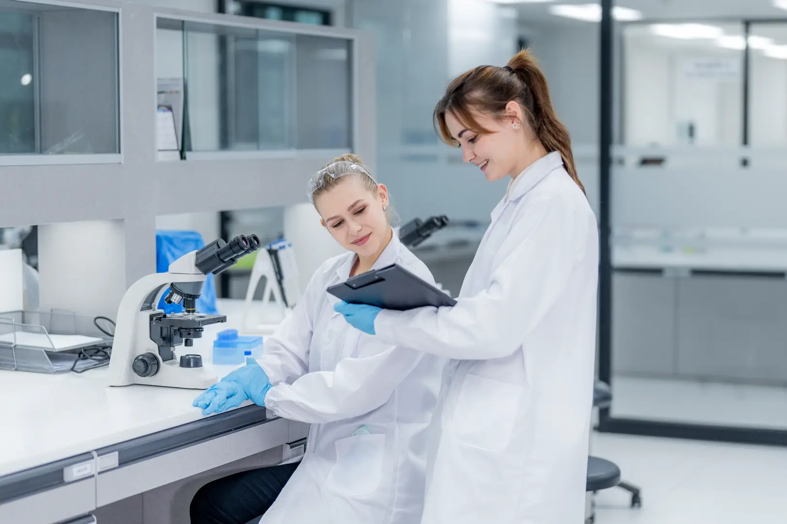 Two researchers in white lab coats reviewing a clipboard beside a microscope in a laboratory.
