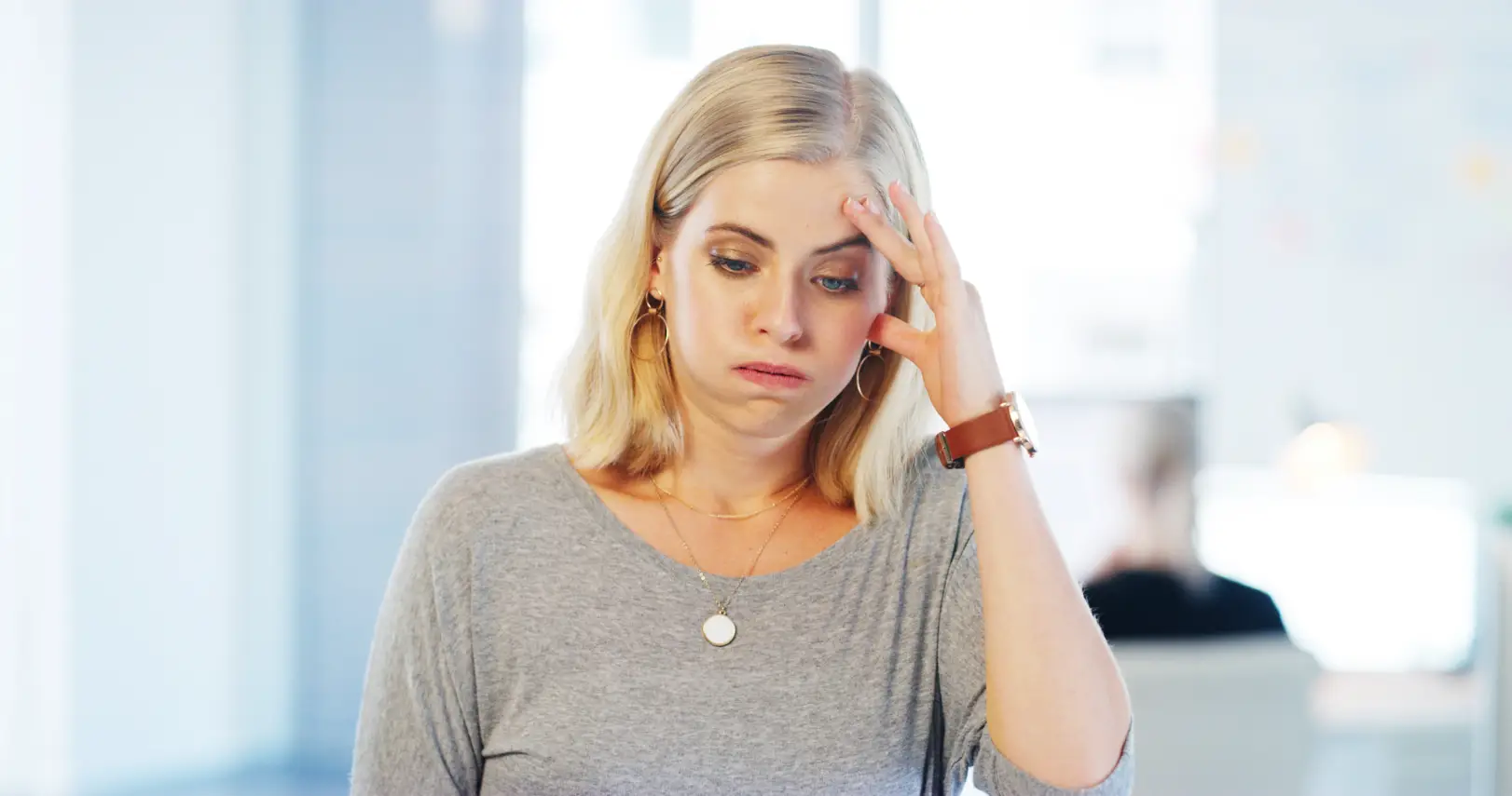 Blonde woman in a gray shirt touching her forehead with one hand while looking downward.